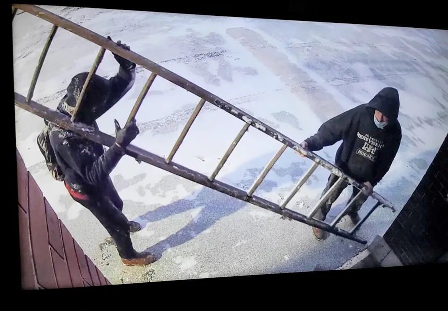 Two men in hoodies carry a ladder on the snowy grounds.
