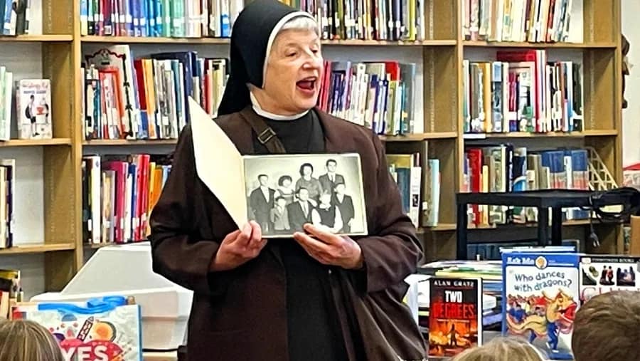 A nun in full habit holds on old photograph. She sits in front of a colorful bookcase.