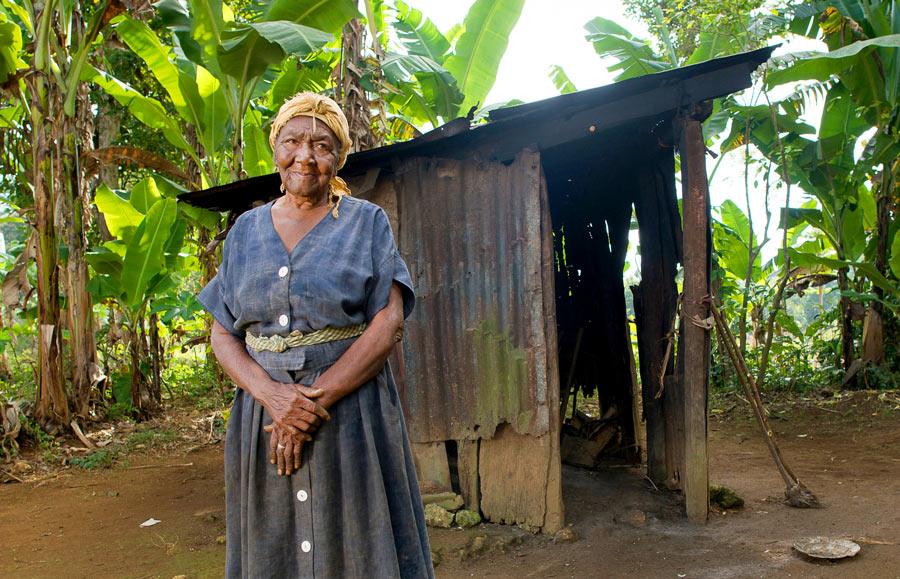 Elderly Haitian woman standing in front of her dilapidated home.